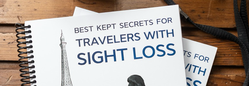 This photo shows two copies of Best Kept Secrets For Travelers With Sight Loss lying on a rich golden-brown-colored wood table. The top book is angled over the bottom one, mostly covering it. Also visible on the wood surface are a half-full cup of coffee and a camera (above the books) and a pen (below the books). The top book, the large-print edition, is coil-bound for ease of reading if a person is using magnification aids. The bottom book is bound normally. The book cover is mainly black and white. In the foreground is a female traveler shown in side view. She is striding purposefully, holding her white cane in front of her in her gloved right hand. The bottom quarter of the cane is bright red. The traveler is dressed in practical, yet attractive clothing: a peacoat of above-the-knee length, a small backpack, and sturdy shoes. The title of the book appears in the upper right part of the cover, with the author’s name and credentials at the bottom. The cover background shows the Eiffel tower, a few roughly sketched trees, and three silhouetted figures.